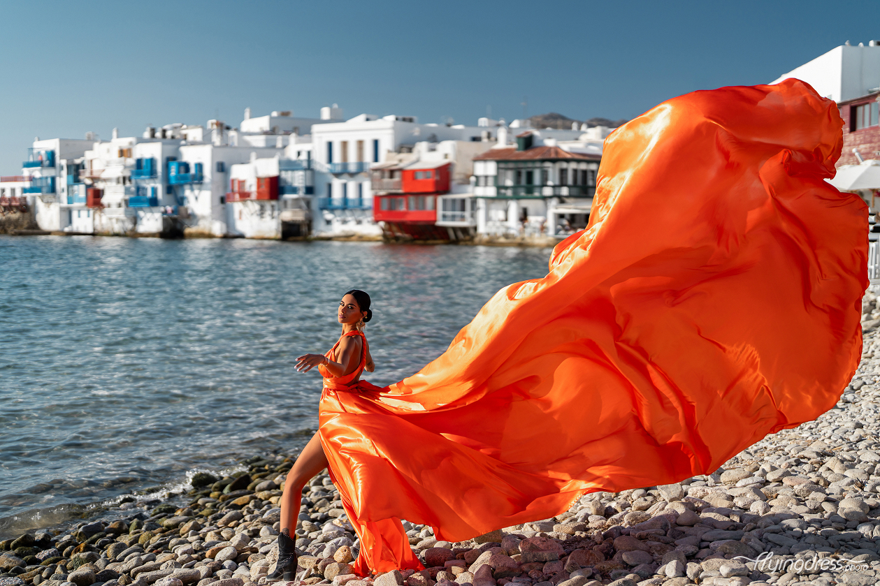 A woman in a striking orange gown poses elegantly on the rocky shoreline of Mykonos' Little Venice, with the vibrant backdrop of the iconic waterfront houses.